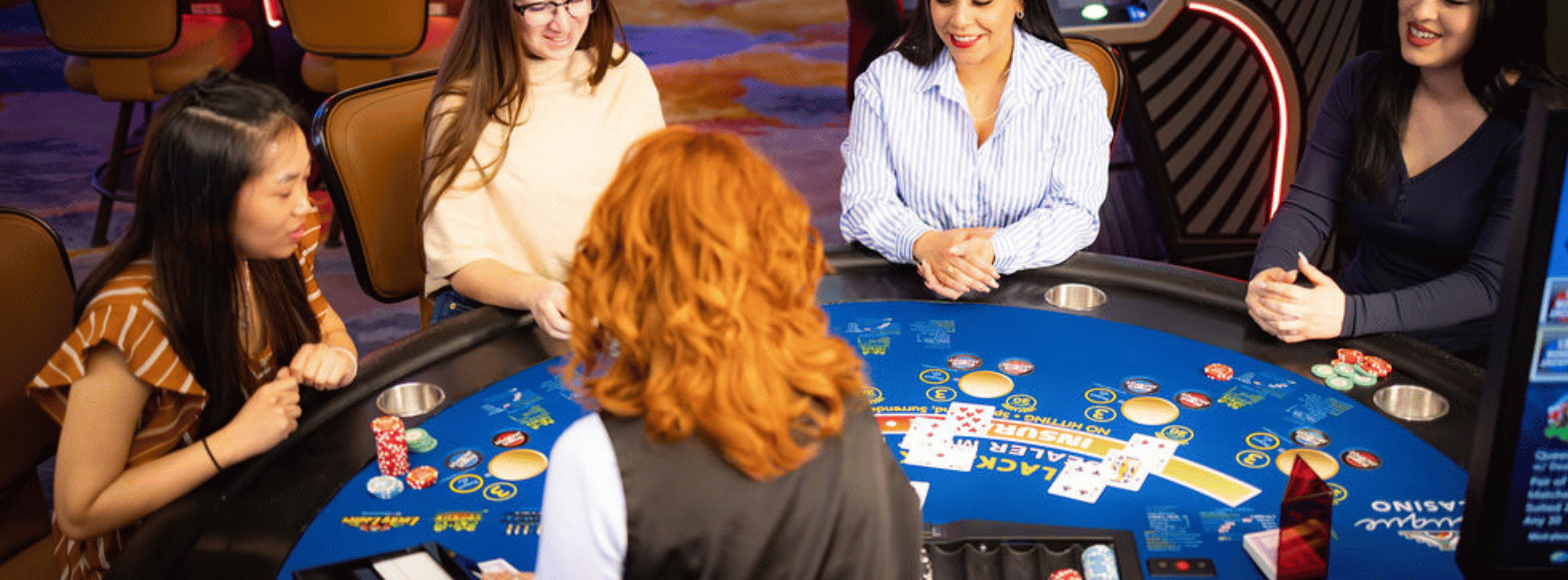 Five women sit around a blue blackjack table, with cards and chip stacks in play at a casino.